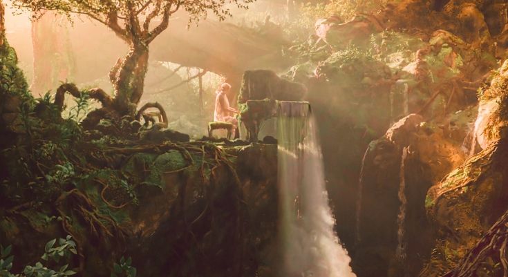 a woman standing on top of a waterfall next to a forest filled with lush green trees