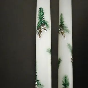 two white candles decorated with pine cones and evergreen needles