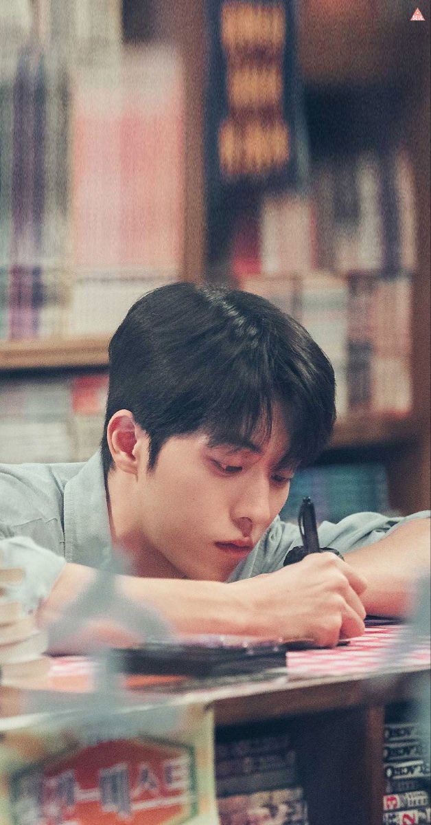 the young man is writing at his desk in front of bookshelves and shelves