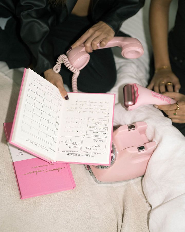 two women are sitting on a bed and one is holding a pink telephone, while the other holds a notebook