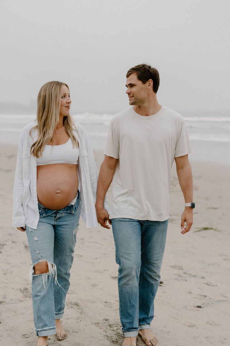 a pregnant couple walking on the beach holding hands