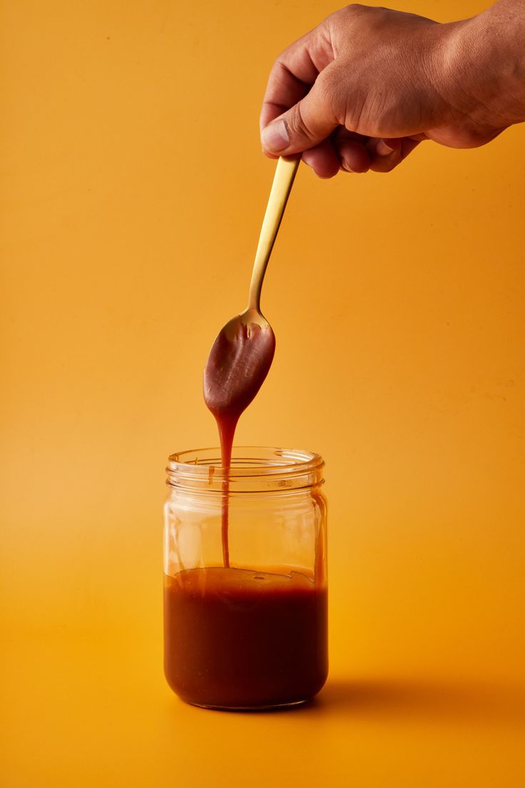 a hand holding a wooden spoon over a jar filled with chocolate sauce on an orange background