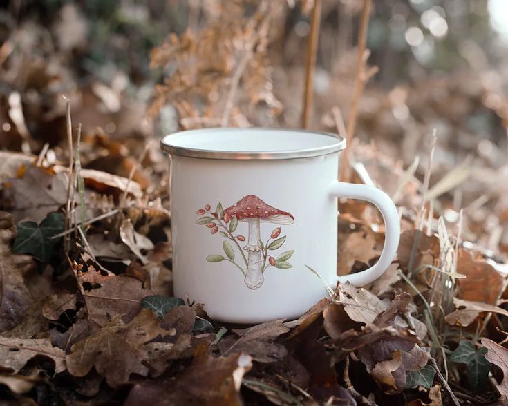 a white coffee cup sitting on top of leaves in the grass with mushrooms painted on it