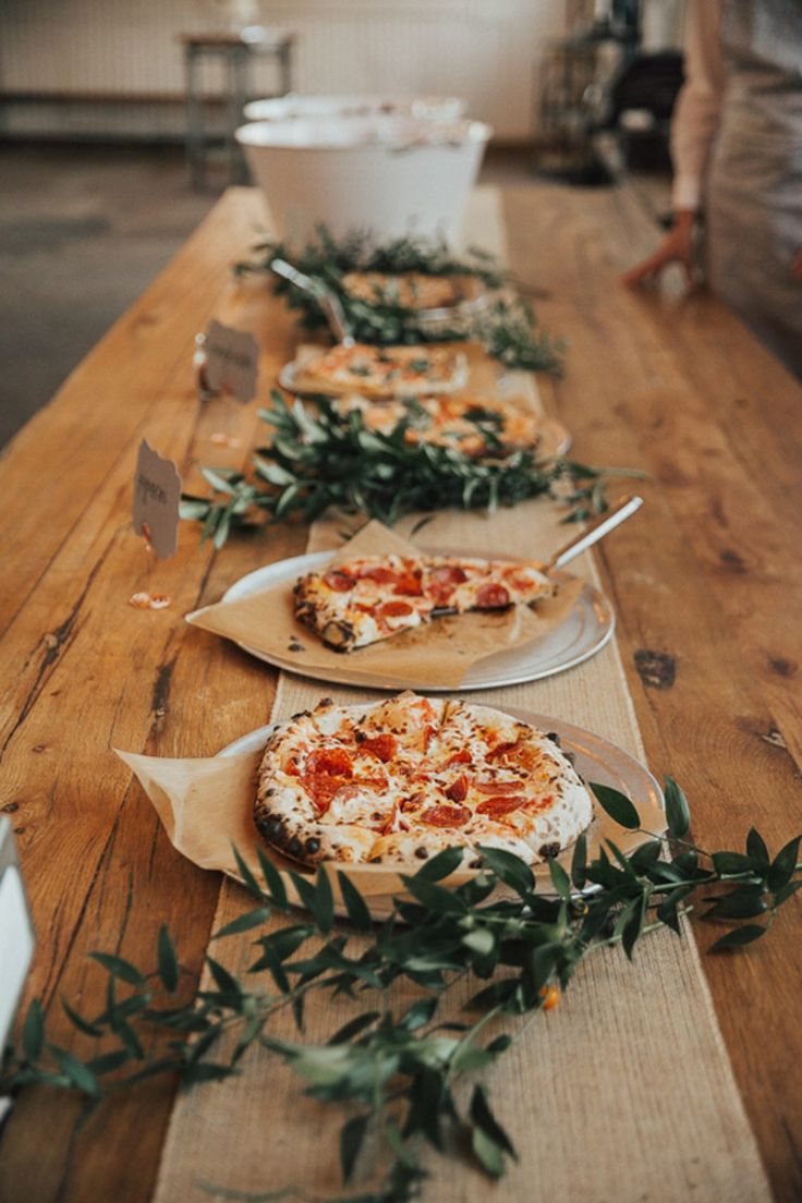 several pizzas on plates lined up on a long table with greenery around them