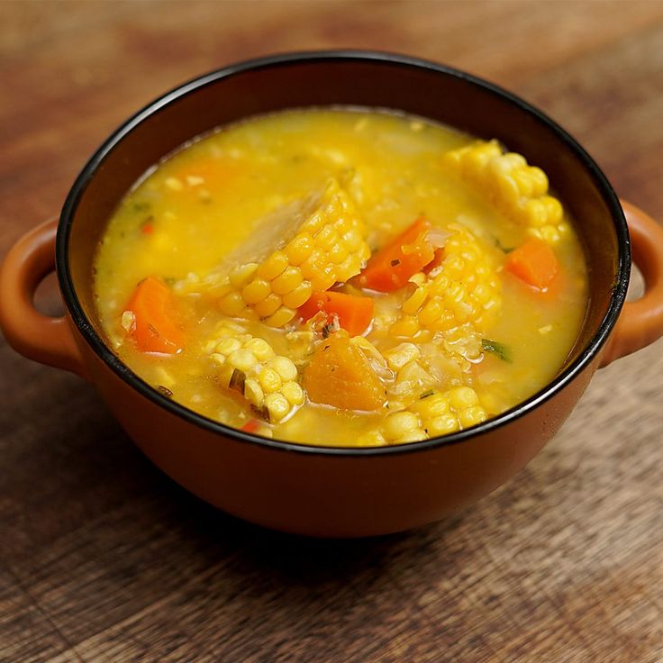 a brown bowl filled with corn and carrots on top of a wooden table next to a spoon