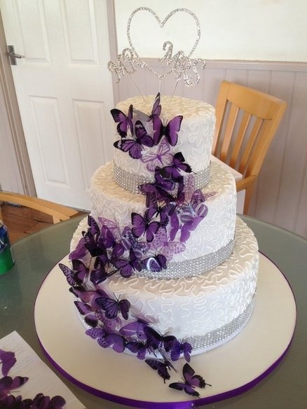 a three tiered white cake with purple flowers on the top and bottom, sitting on a table