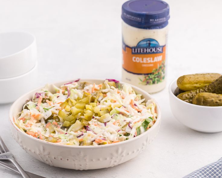 a white bowl filled with coleslaw and pickles next to a container of pickles