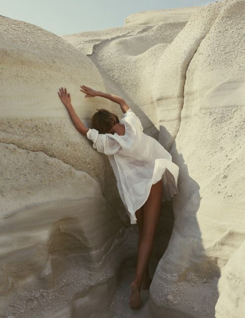 a woman in white shirt and skirt leaning against rock formation with her arms outstretched out