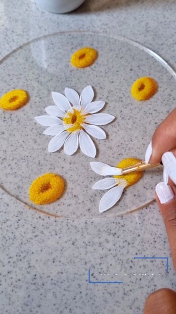 a person is painting flowers on a glass plate with yellow and white dots in the center