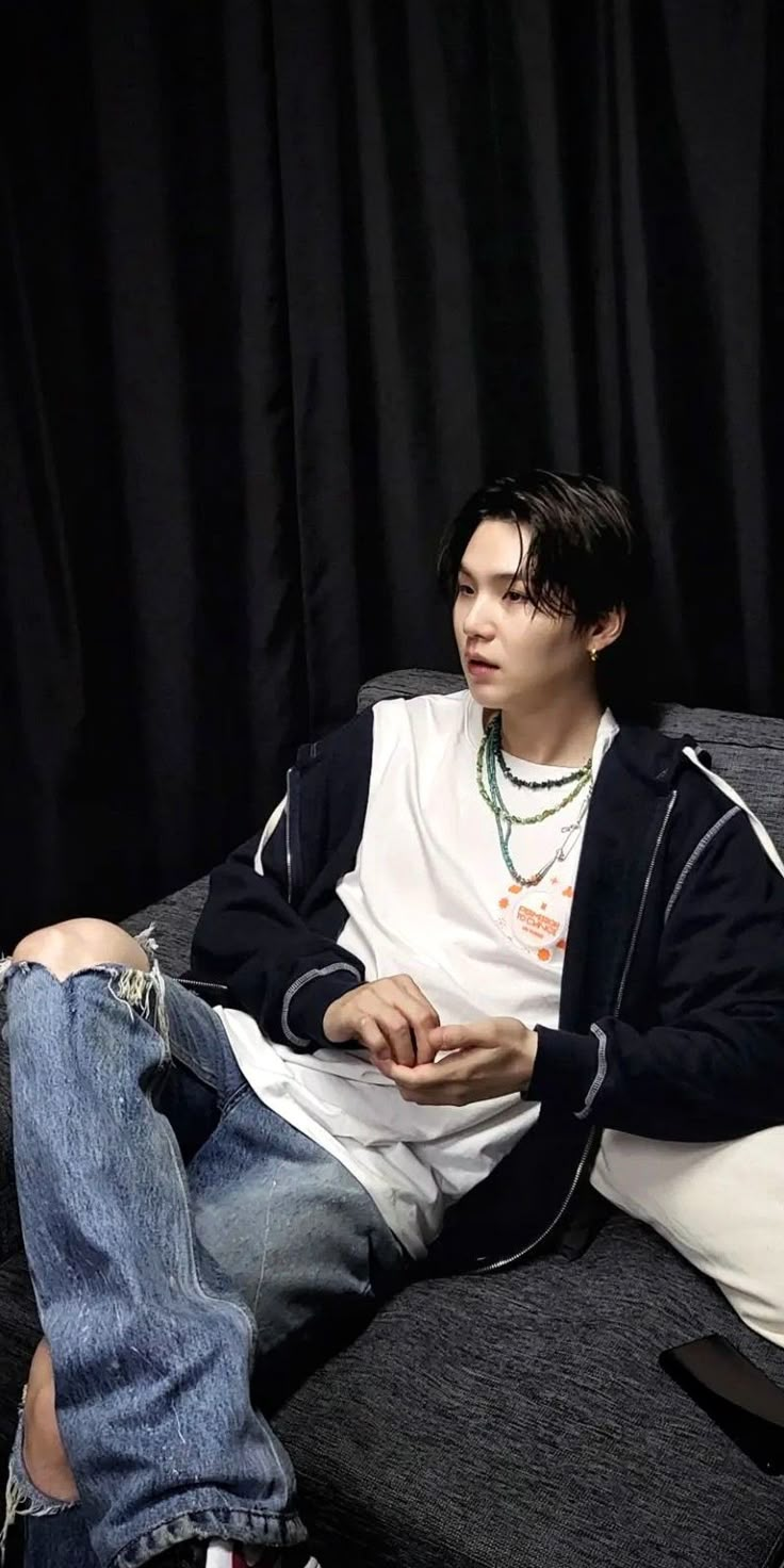 a young man sitting on top of a couch next to a black curtain and wearing a white shirt