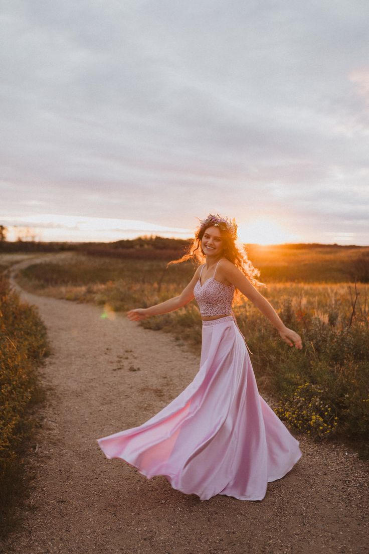 a woman in a pink dress is walking down a dirt road at sunset with her arms outstretched