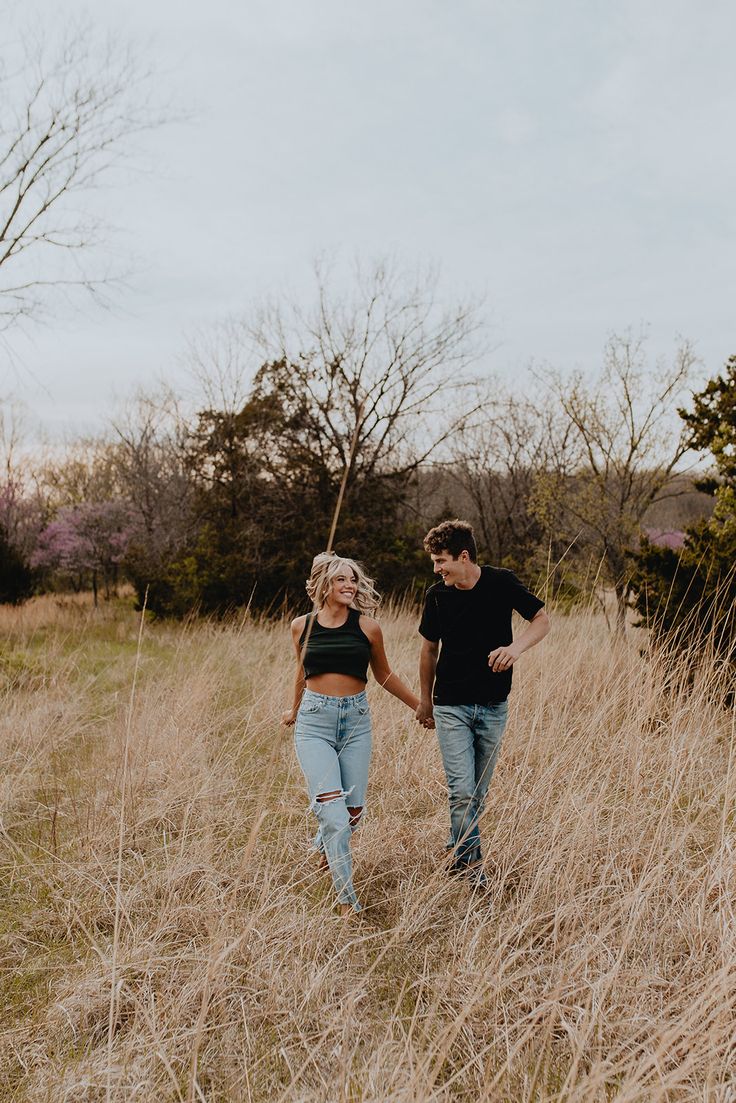 a man and woman holding hands walking through tall grass in an open field with bare trees