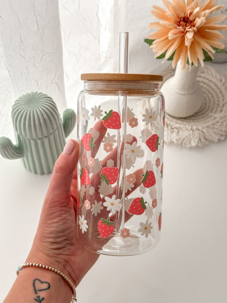 a person holding up a glass jar with strawberries and flowers on the inside, in front of a cactus plant