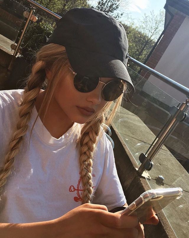 a girl with long braids is looking at her cell phone while wearing a hat and sunglasses