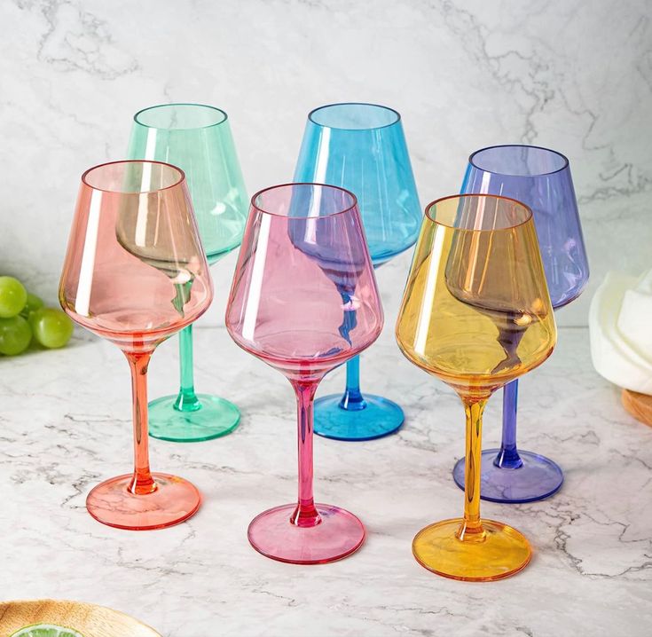 several different colored wine glasses sitting next to each other on a marble counter top with grapes in the background