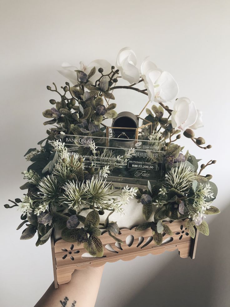 an arrangement of flowers and greenery is displayed on a wooden shelf in front of a white wall