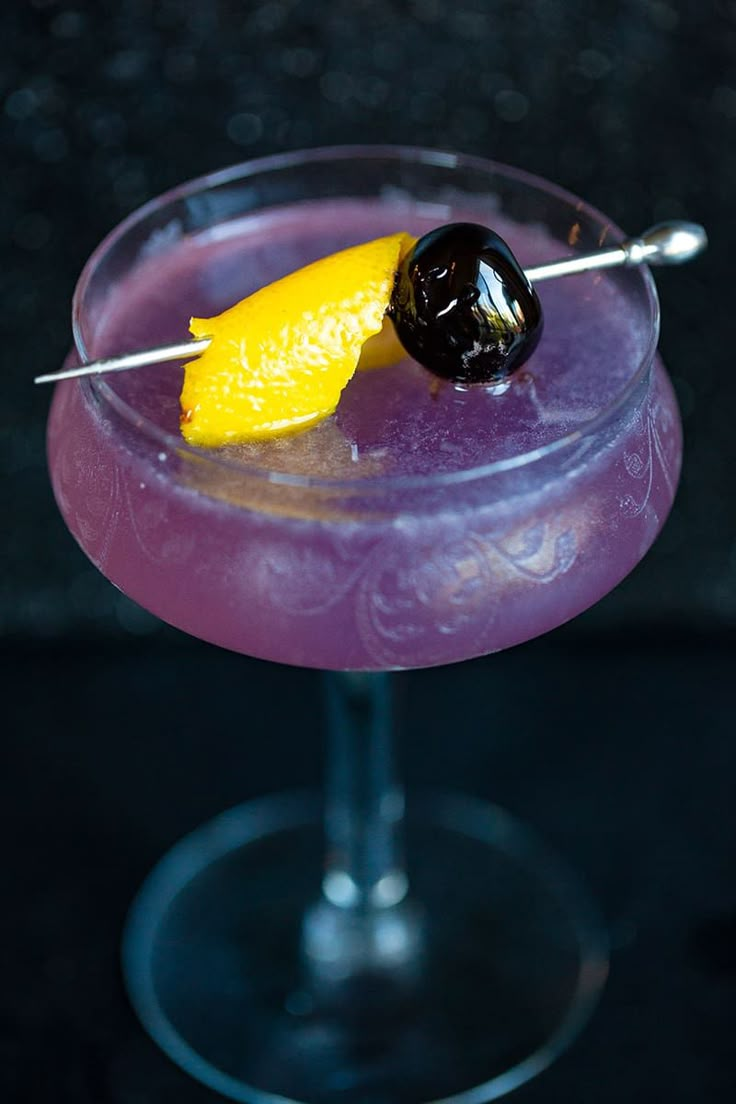 a purple drink with a lemon on the rim and garnished with black olives