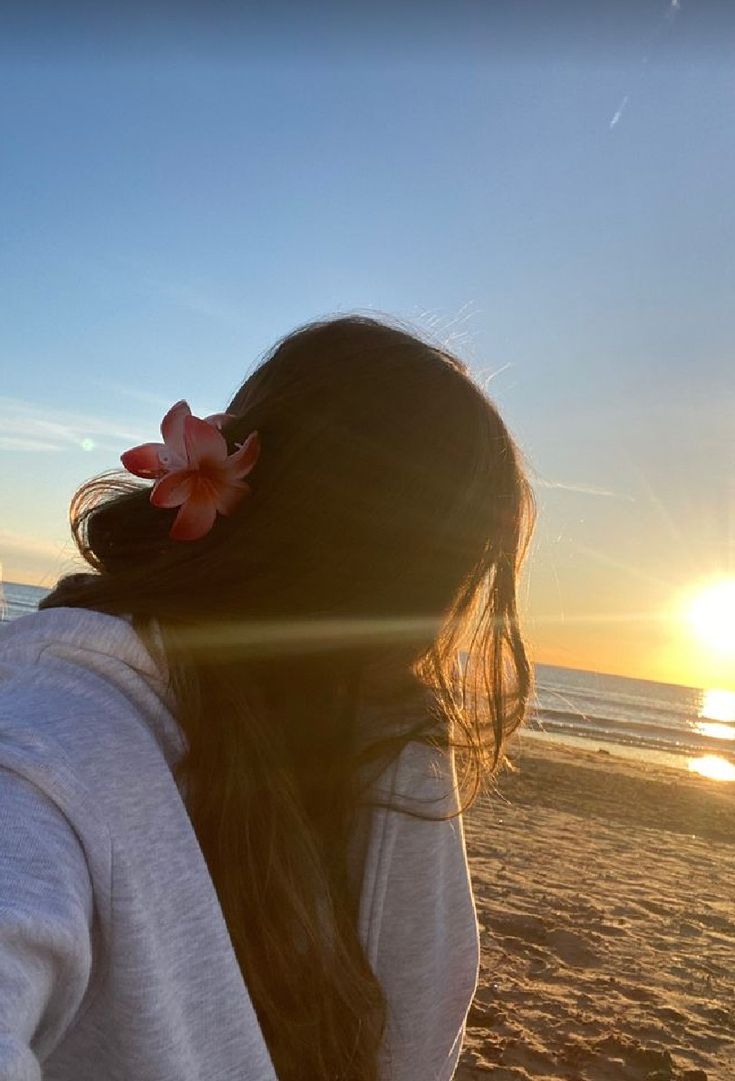 a woman standing on top of a sandy beach next to the ocean with a pink flower in her hair