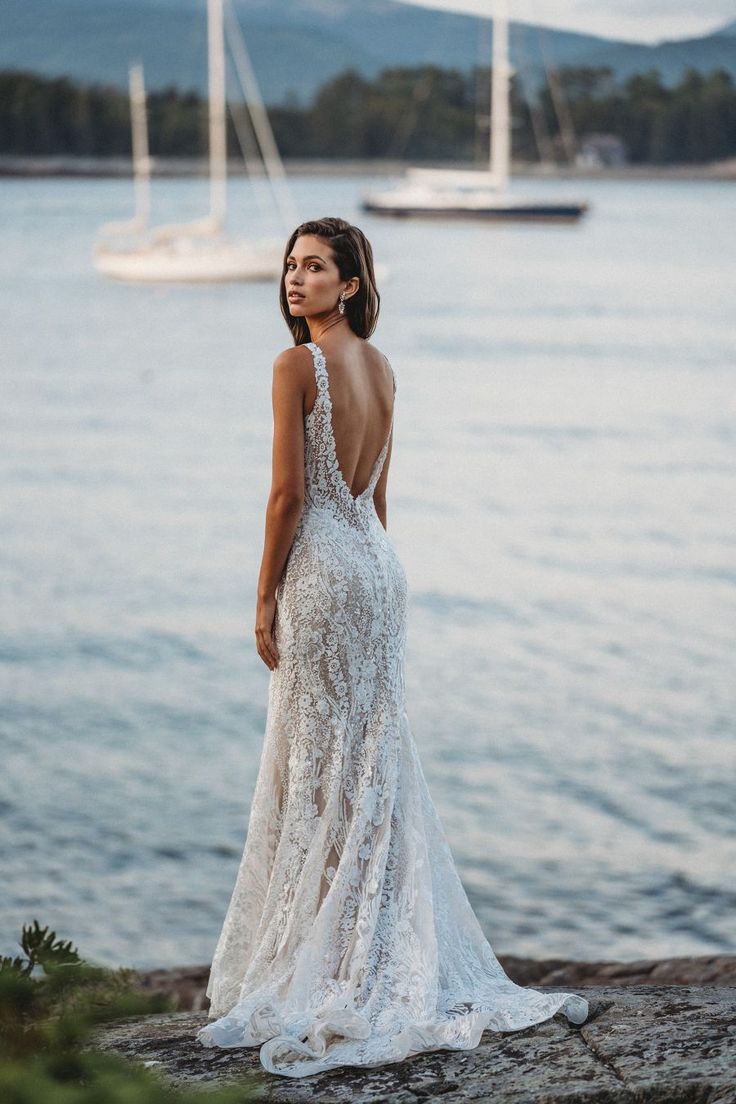 a woman in a wedding dress standing on the shore looking out at the water and yachts