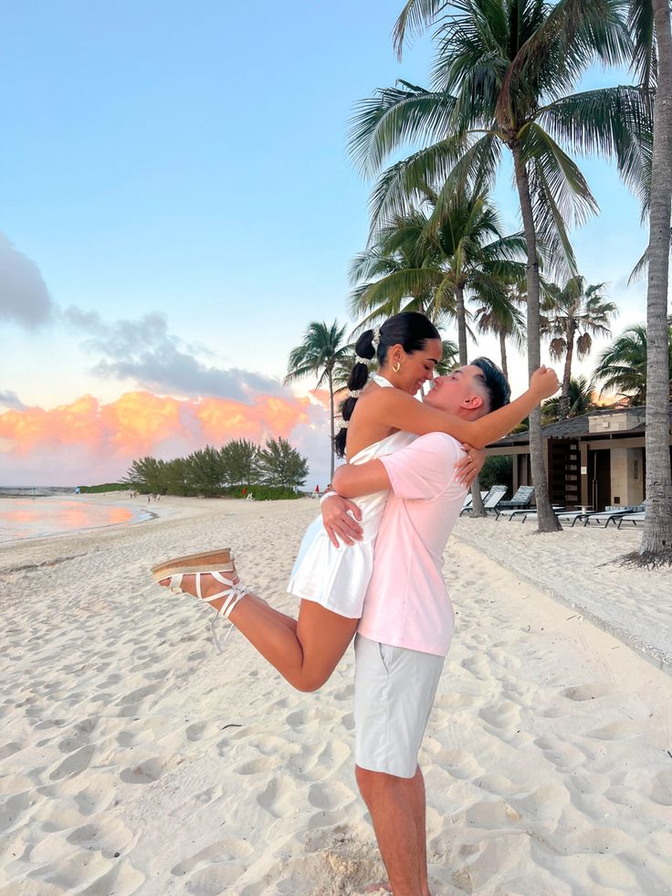 a man and woman are hugging on the beach with palm trees in the background as the sun sets