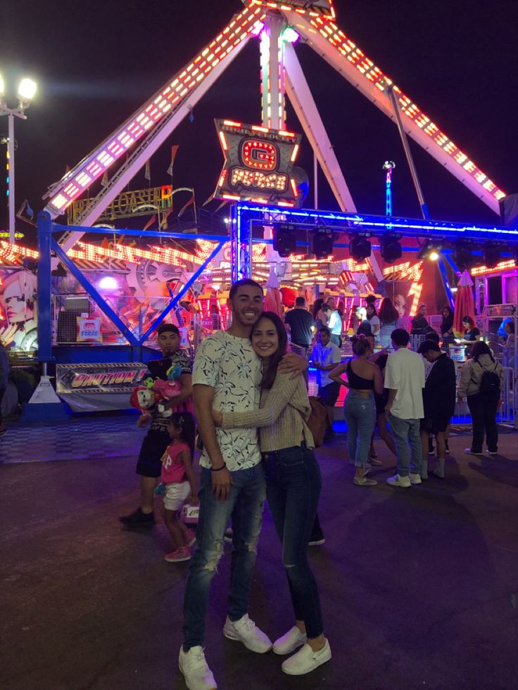 a man and woman standing in front of a carnival ride at night with lights on