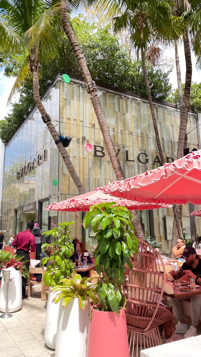 people are sitting at tables under umbrellas in front of a building with palm trees