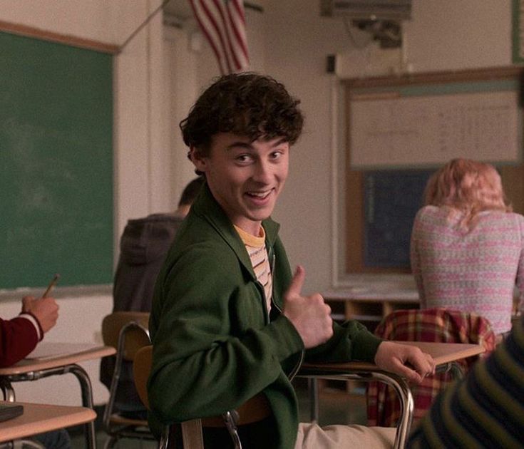 two young men sitting at desks in a classroom with one giving the thumbs up