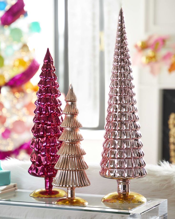 three small christmas trees on a table in front of a window with colorful decorations behind them