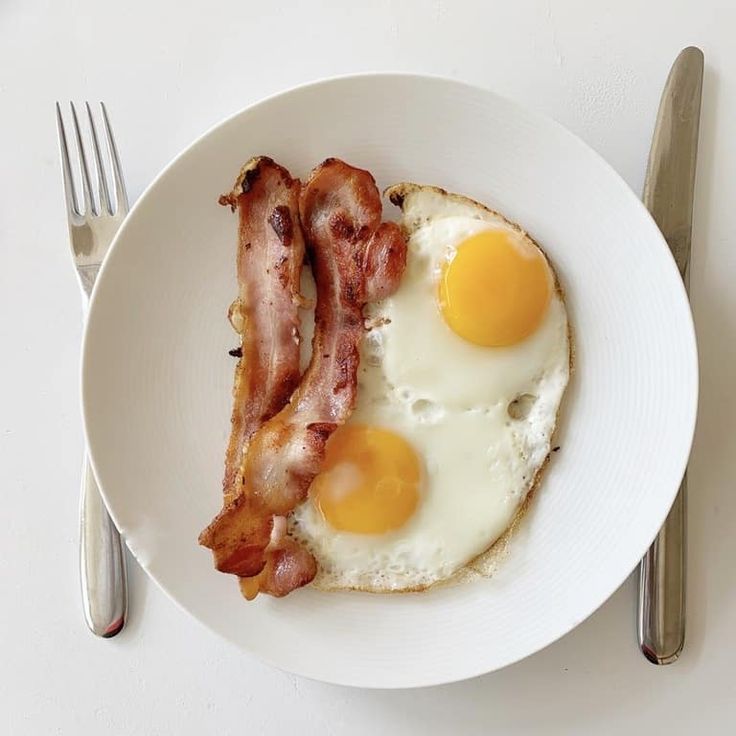 two fried eggs and bacon on a white plate with silverware next to the plate