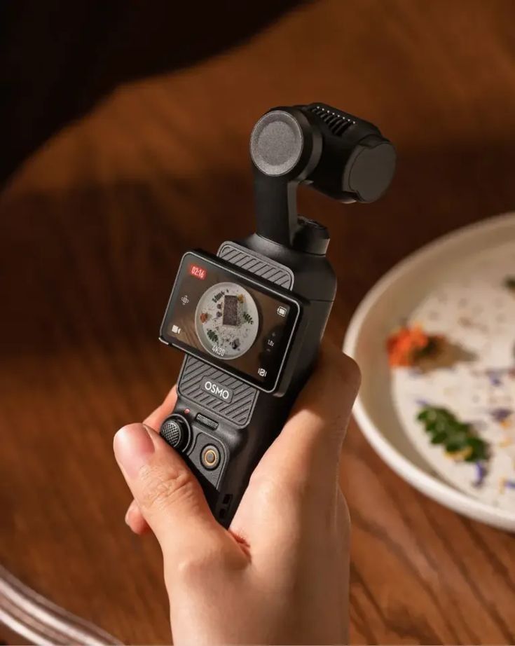 a person holding a camera in their hand on top of a wooden table next to a plate