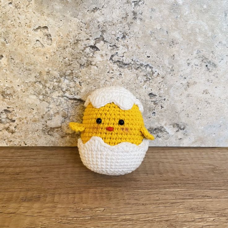 a small crocheted chicken sitting on top of a wooden table next to a stone wall