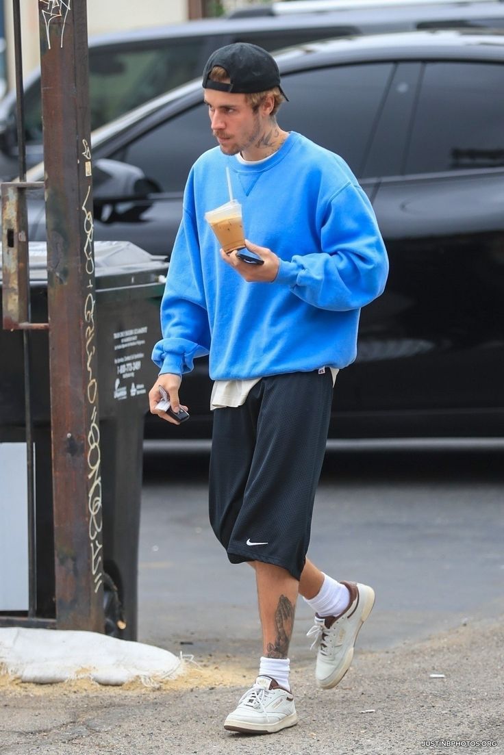 a man walking down the street while wearing a blue sweatshirt and black sweatpants, holding a drink in his hand