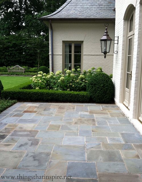 an outdoor patio with stone pavers and shrubbery in the foreground, next to a white brick house