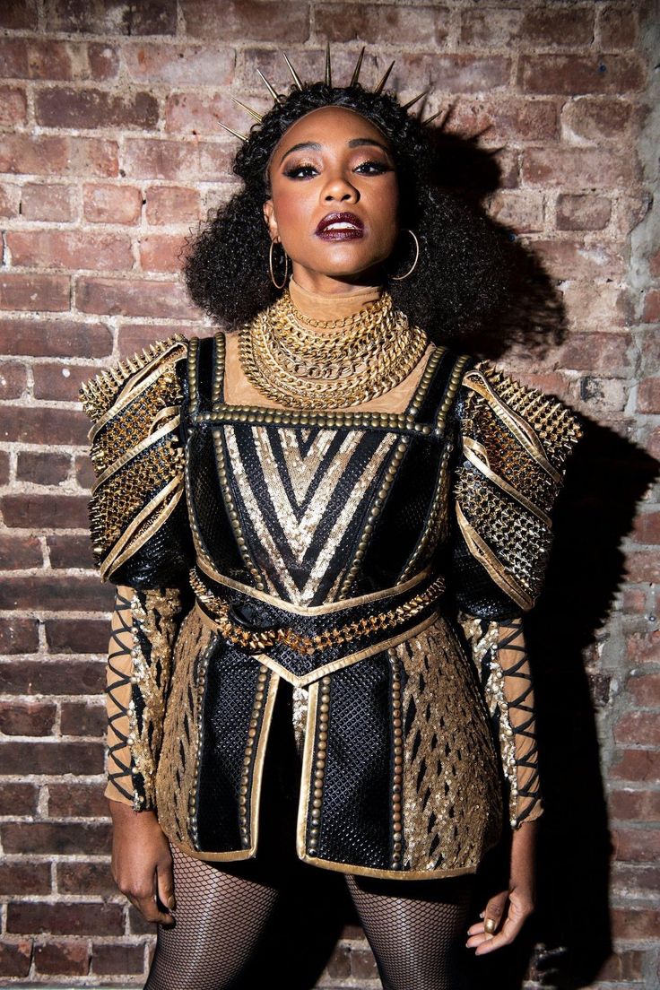 a woman in black and gold outfit standing next to a brick wall with her hands on her hips