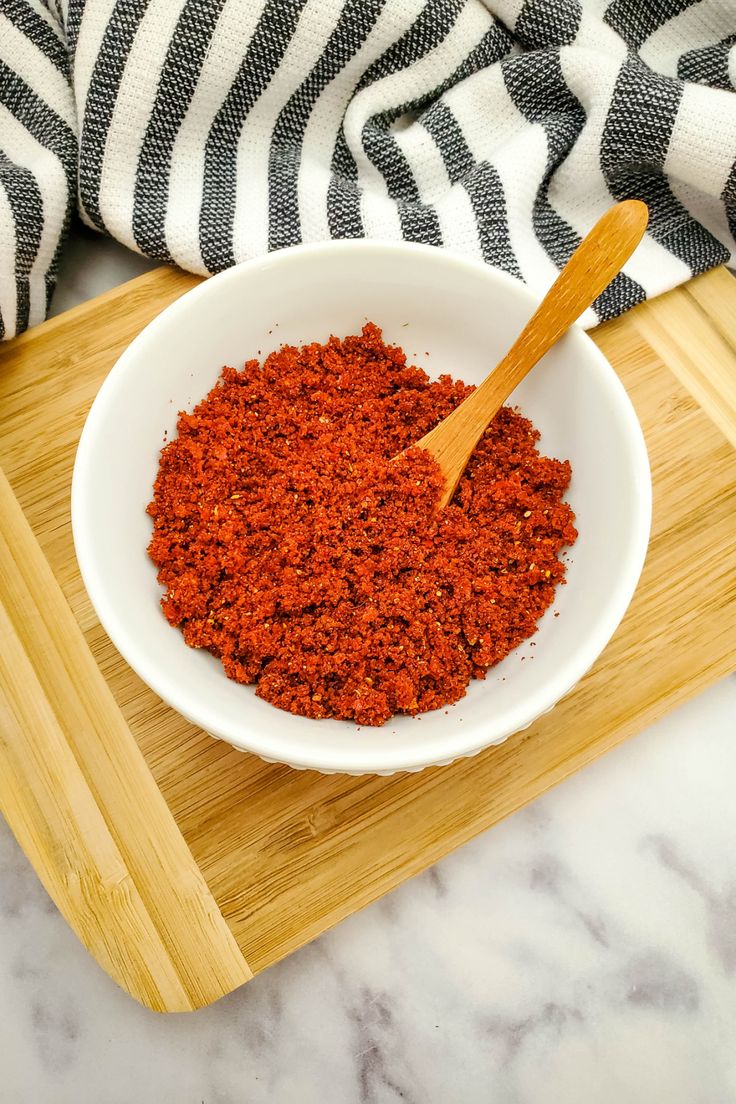 a wooden spoon in a white bowl filled with ground chili on a cutting board next to a black and white towel