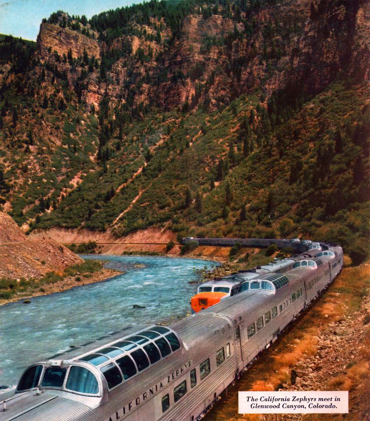 a train traveling down tracks next to a river and mountain side covered in greenery