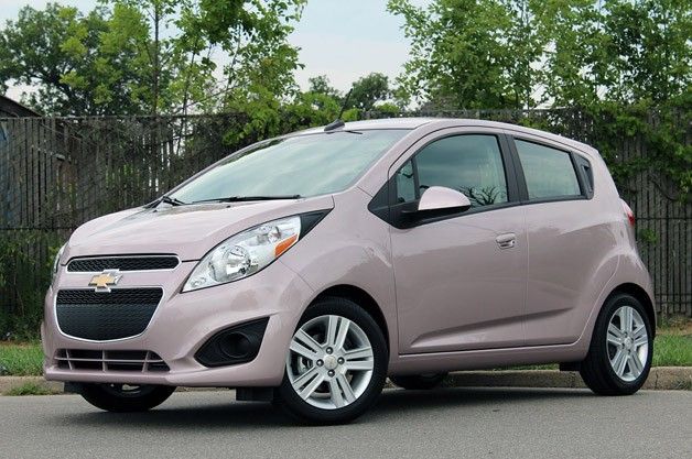 a pink chevrolet spark is parked on the side of the road in front of a fence