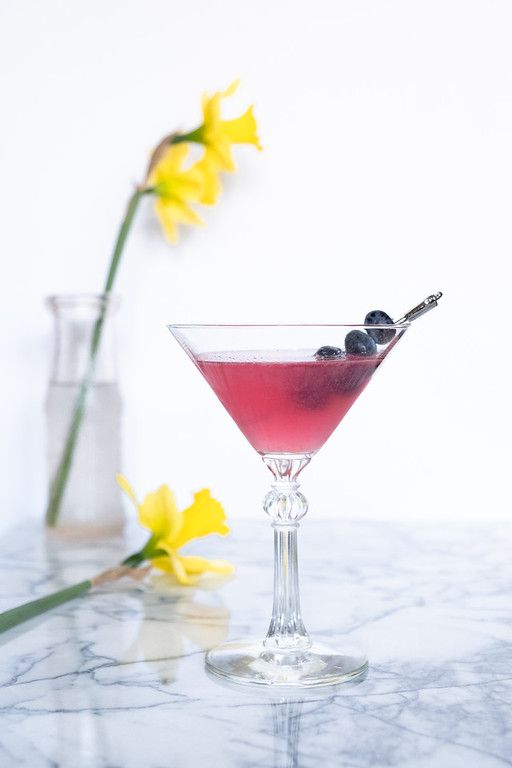 a glass filled with a red liquid and some black olives on the rim next to yellow daffodils