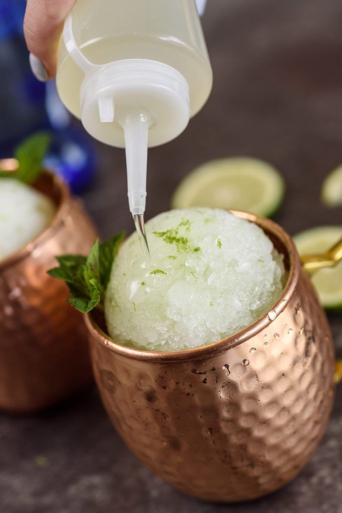 a person pouring sugar into a copper cup filled with ice and lime slices on the side