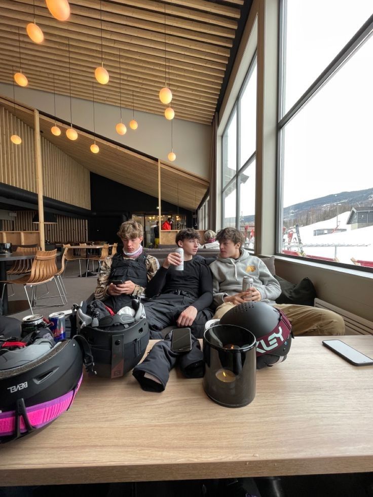 three people sitting on a table with luggage and helmets in front of them, all looking at their cell phones