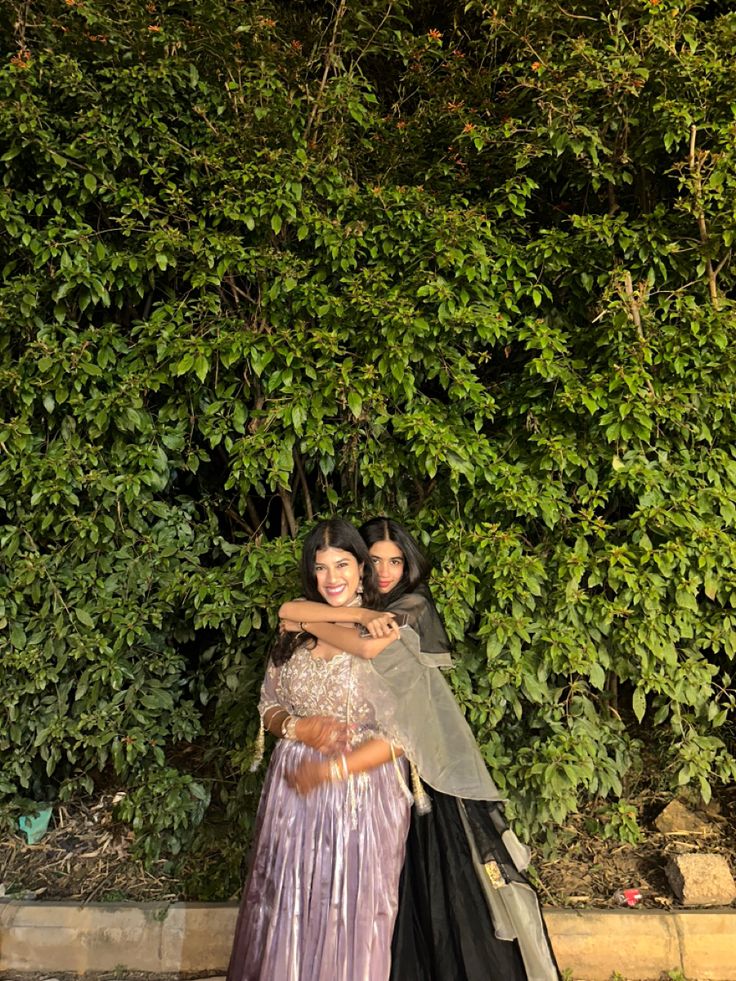 two women standing next to each other in front of a tree and bushes at night