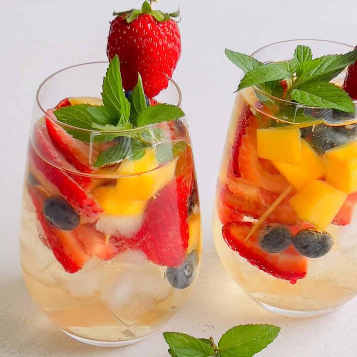 two glasses filled with fruit and ice on top of a white table next to each other