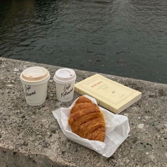 there is a croissant, coffee and book on the ledge by the water