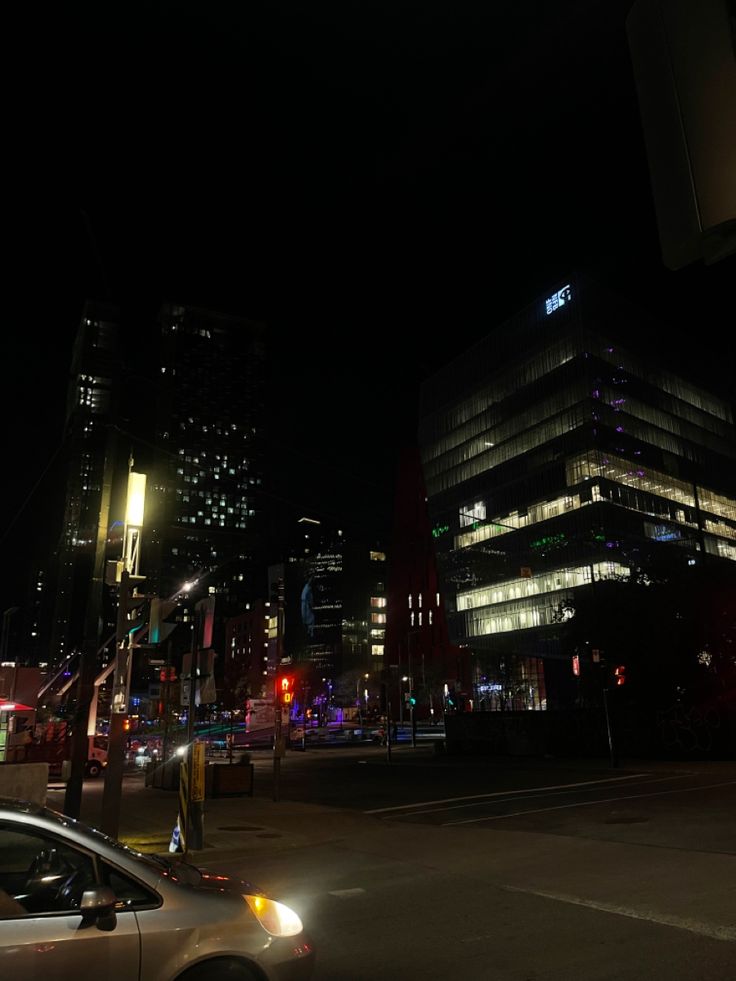 a car is driving down the street at night in front of tall buildings and skyscrapers