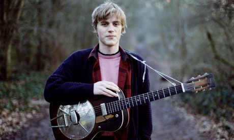 a young man holding an acoustic guitar in his hands while standing on a path through the woods