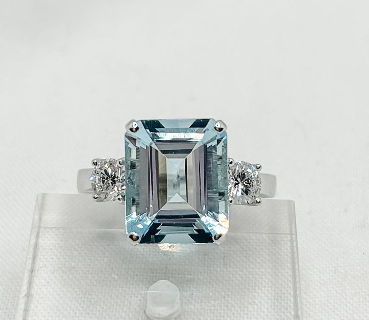 18ct White Gold Aquamarine & Diamond Ring Description This stunning 18ct white gold ring is a masterpiece of elegance, featuring a beautifully cut emerald aquamarine at its center. The aquamarine, with its captivating light blue hue and loupe-clean clarity, is complemented by two dazzling round brilliant-cut diamonds. Each diamond is securely set in four claws and showcases exceptional sparkle, making this ring an exquisite choice for someone with a refined taste for fine jewelry. The design is Aquamarine Brilliant Cut Diamond Ring For Formal Occasions, Formal Aquamarine Diamond Ring With Brilliant Cut, Luxury Gia Certified Aquamarine Ring, Elegant Gia Certified Blue Topaz Diamond Ring, Formal Emerald-cut Aquamarine Diamond Ring, Formal Emerald Cut Aquamarine Diamond Ring, Elegant Gia Certified Aquamarine Rings, Elegant White Aquamarine Rings, Elegant Aquamarine Sapphire Ring With Brilliant Cut