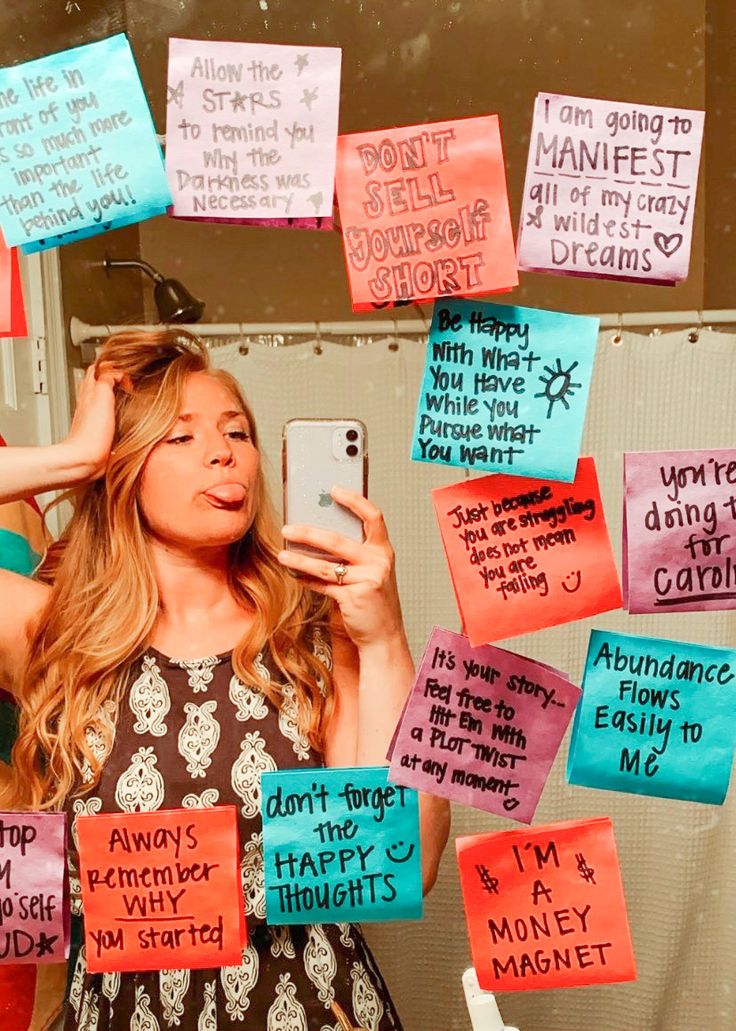 a woman standing in front of a mirror holding a cell phone and surrounded by post it notes