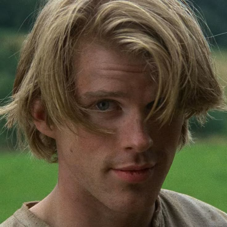 a young man with blonde hair and blue eyes looks at the camera while standing in front of a green field