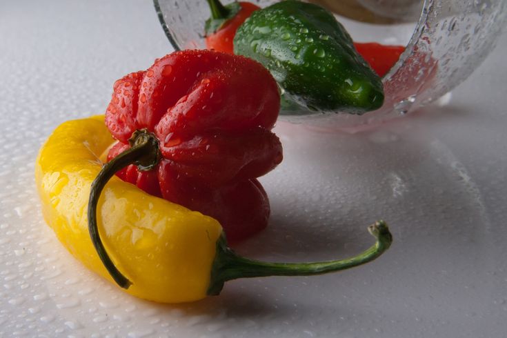 three peppers sitting on top of a table next to a glass bowl filled with red, yellow and green peppers