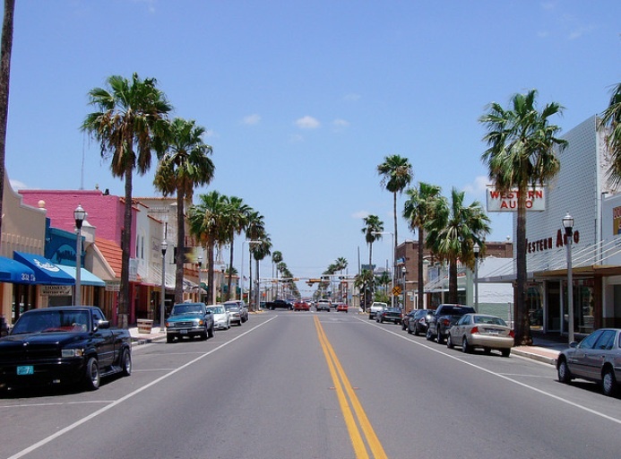 cars are driving down the street in front of shops and palm trees on both sides